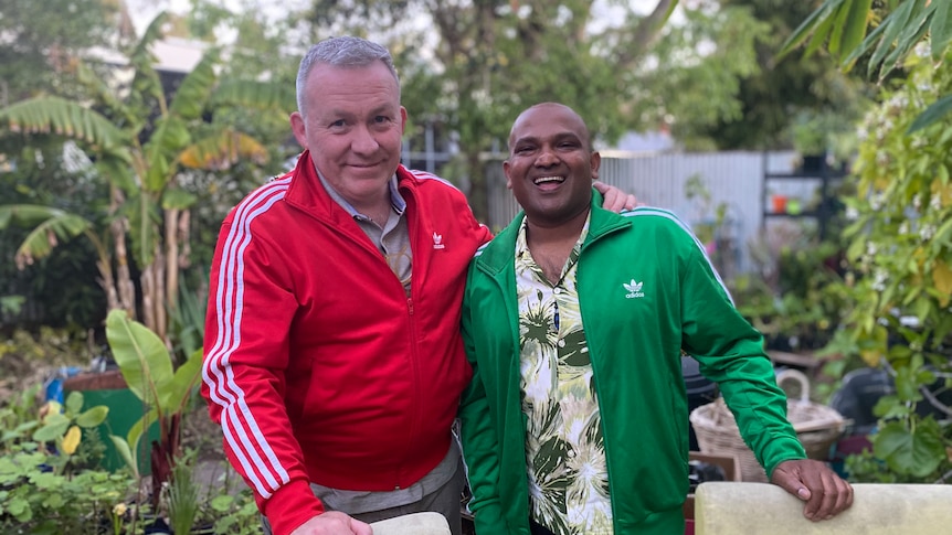 Daniel, wearing a red jacket, and Devanesan, a green jacket, smile at the camera, with a lush garden behind them.