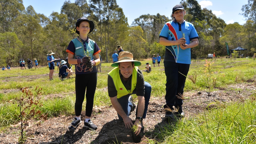 A man in a hi-vis vest plants a seedling while two school students stand next to him.