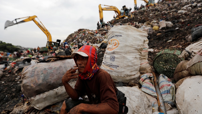 A scavenger smokes a cigarette sitting on rubbish at a landfill. 