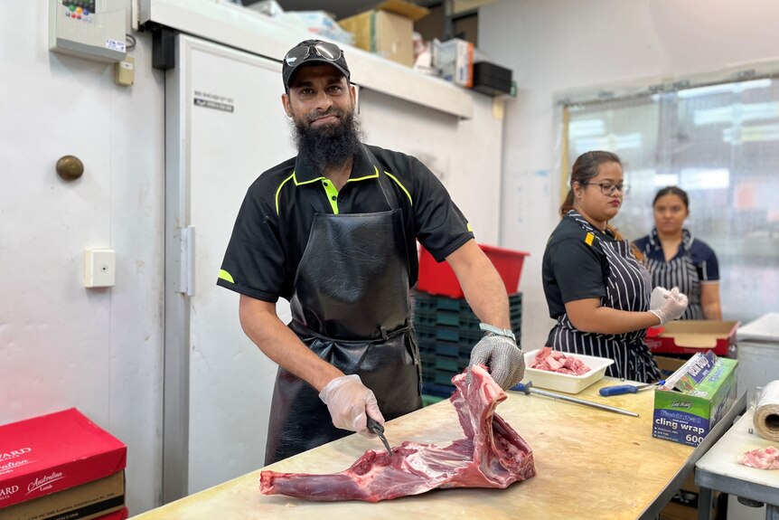 A man wearing an apron slices a large piece of meat on a table, inside a commercial kitchen.