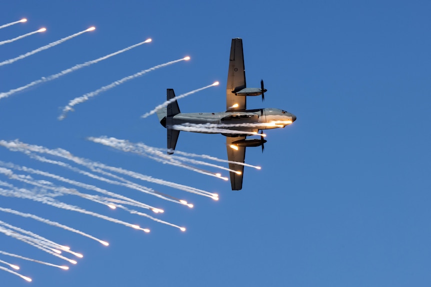 A plane flies against the blue sky as multiple flares shoot out around it.