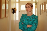 A woman with blonde hair wearing a green blouse stands centred with arms crossed in a hallway, serious facial expression