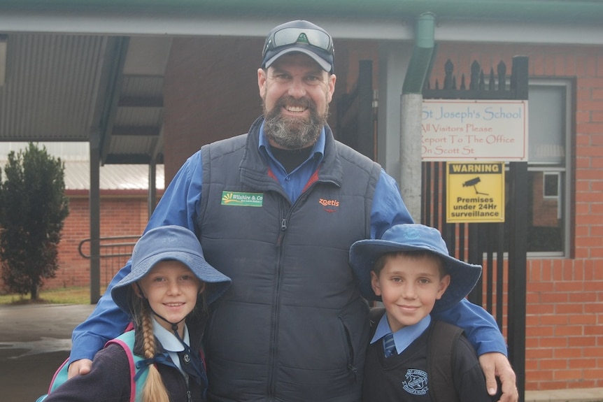 A man stands in front of a school, with an arm around his two children.