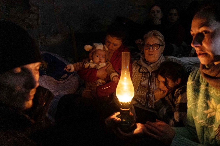 A group of people, including children, huddle around the light of a globe in a dark room.