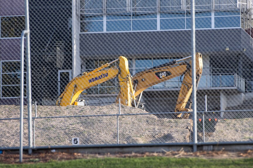 Excavator on grounds of a school, seen behind a fence.
