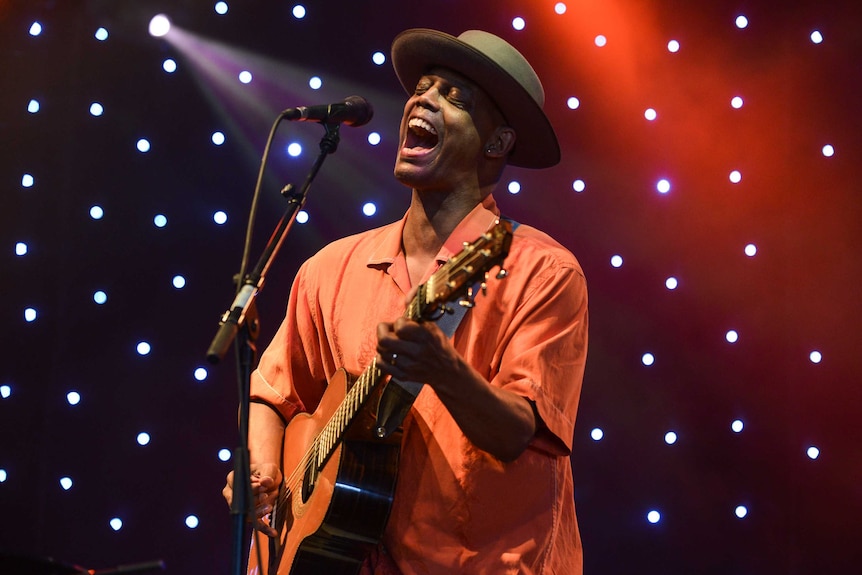 Eric Bibb performs on stage with a guitar. He's wearing an orange shirt, green hat and singing with his eyes closed.