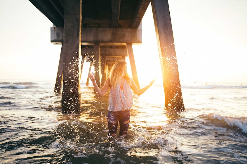 Woman splashes around in the ocean wearing jeans while the sun sets to depict theories about denim and jeans.