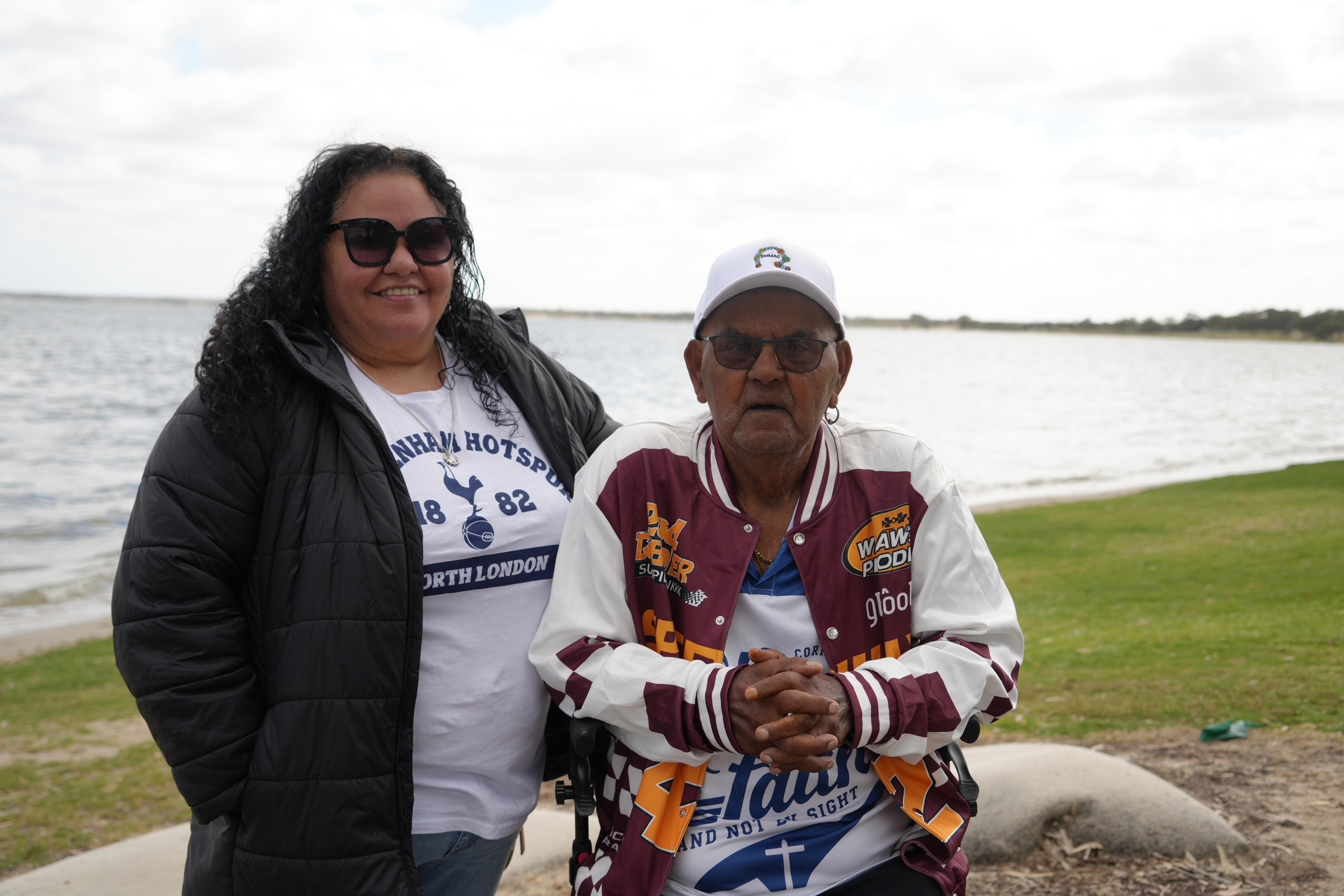 An Aboriginal woman wearing sunglasses standing next to an older Aboriginal man on the shore of a large lake.