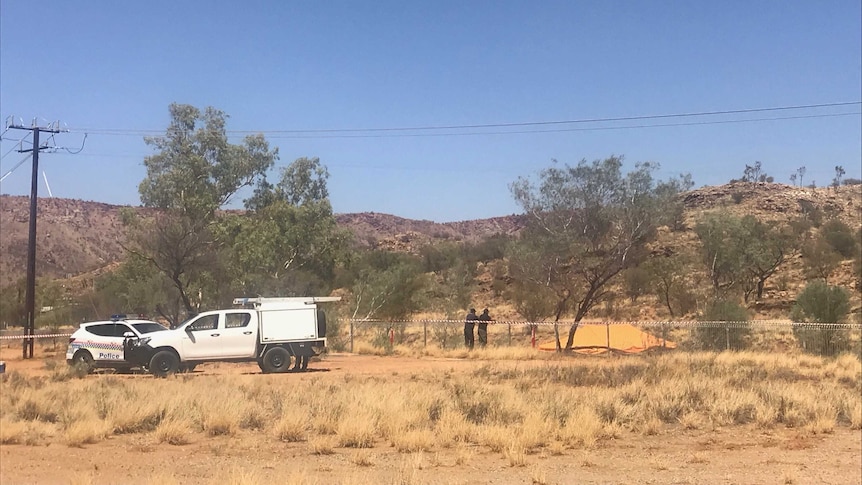 Police vehicles parked near a dirt road in Alice Springs. Two people lean on a fence.