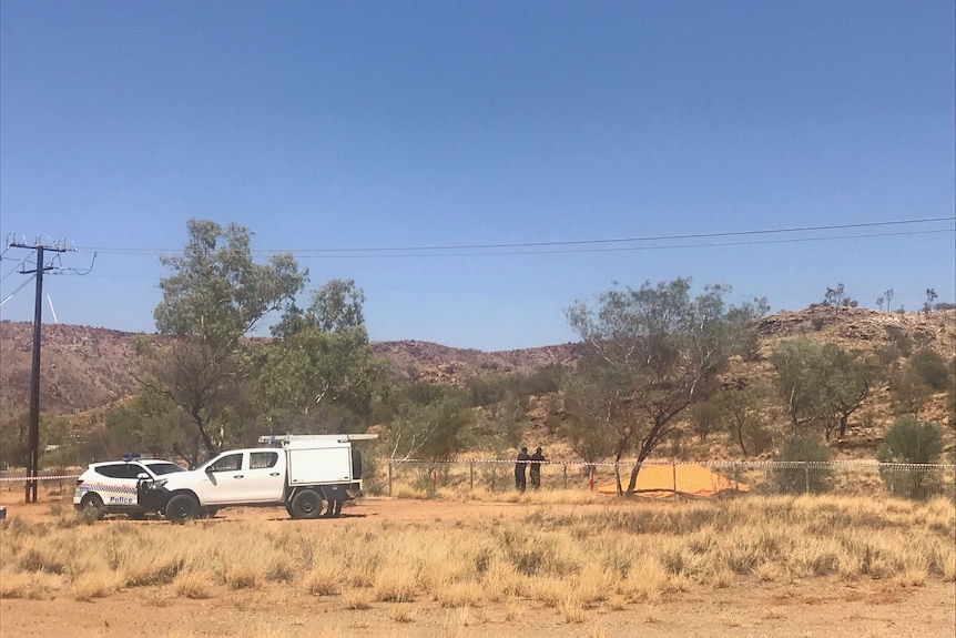 Police vehicles parked near a dirt road in Alice Springs. Two people lean on a fence.
