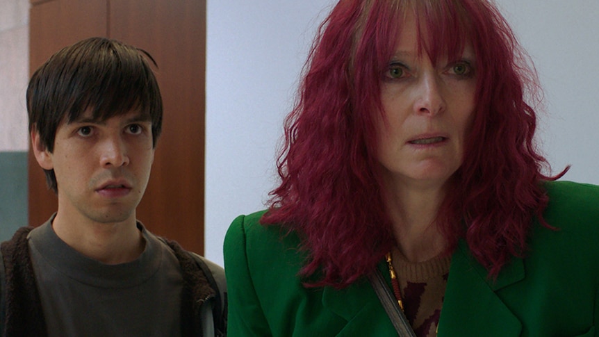 A woman with dyed purple hair looks distressed. She is wearing a forest green blazer. Behind her, a man in a hoodie.