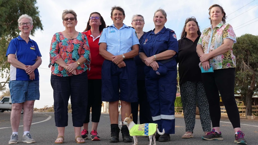 Eight women stand in the middle of a street in a small town and look down at the camera from a low angle.