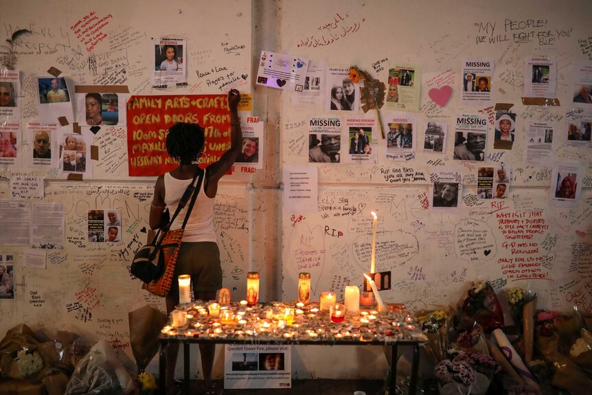 A woman writes on a wall covered with tributes to and pictures of the victims. There's also a table covered in candles.
