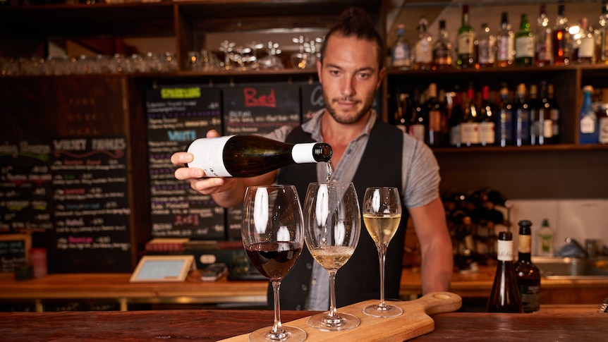 A man behind a bar pours wine into three glasses.