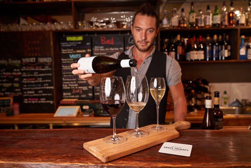 A man behind a bar pours wine into three glasses.