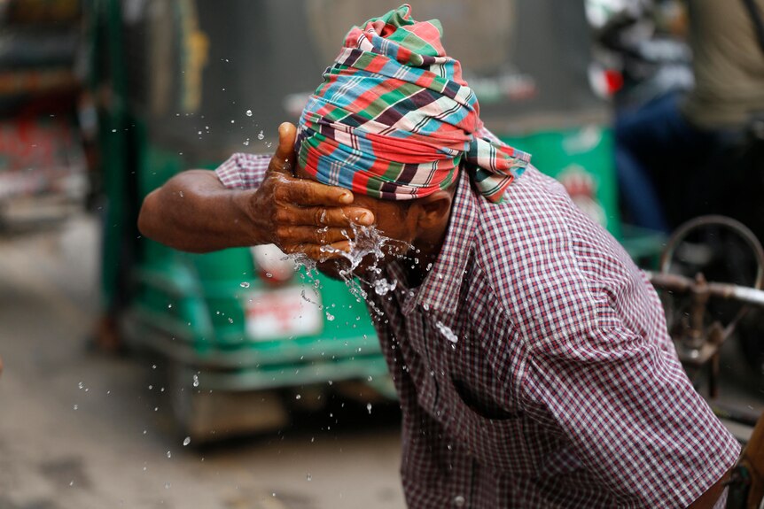 A man in a shortsleeve shirt and headscarf splashes water on his face with his right hand.
