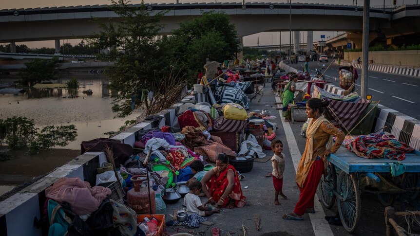 A woman and child stand on a roadside where piles of bags, clothes and other belongings line the edge. 