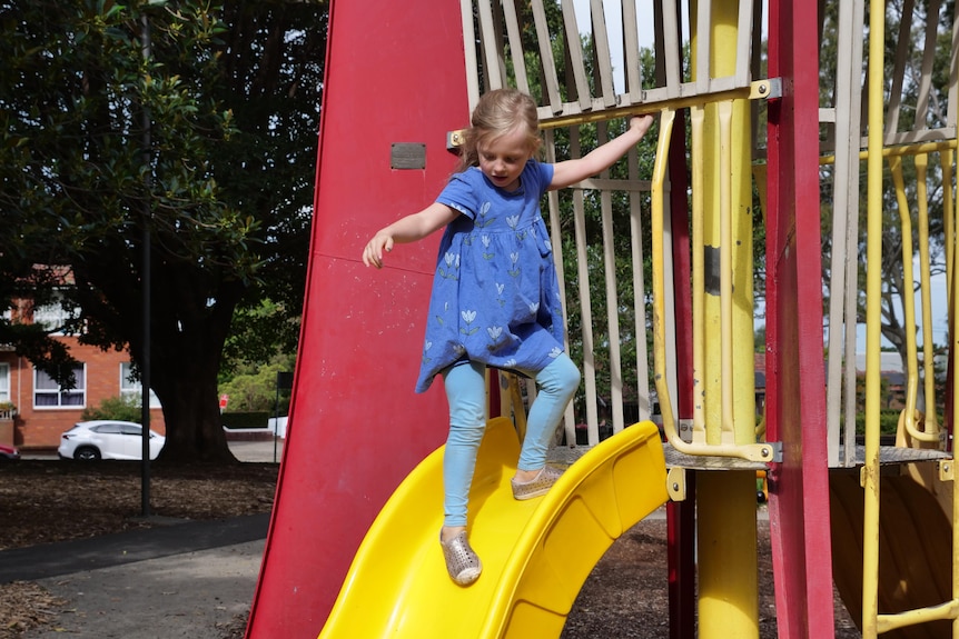 A young girl walks down a yellow slide while holding onto a safety bar on play equipment shaped like a rocket