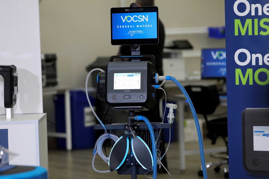 You view a ventilator on display in a manufacturing room with the words 'Vocsn' and 'General Motors' written on its front.