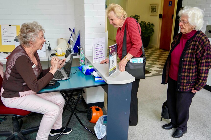 A woman sits behind a desk with a menu and covid check in signs displayed, and two women stand in line