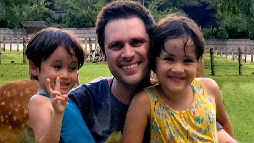 Scott McIntyre hugs his kids while posing for a photo. One gives a peace sign. There is a deer behind them.