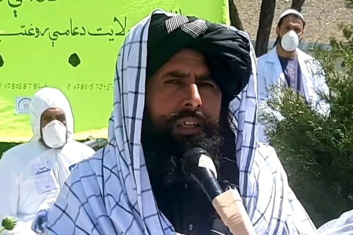 A man with a black beard and a black turban draped in a checked cloth speaks into the microphone.