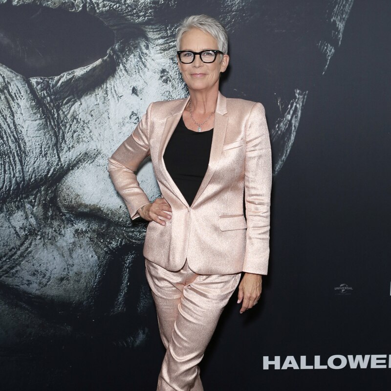 Jamie Lee Curtis on red carpet, in shiny pale pink pants suit over black top, short grey hair and black-rimmed glasses.