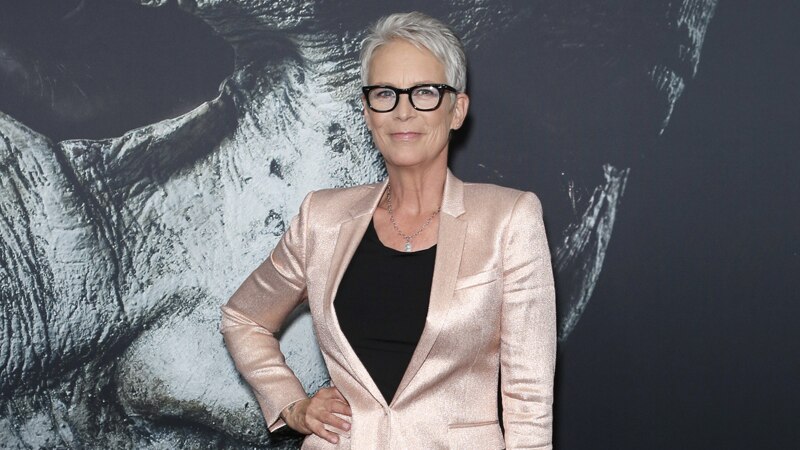 Jamie Lee Curtis on red carpet, in shiny pale pink pants suit over black top, short grey hair and black-rimmed glasses.