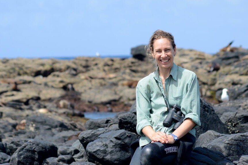 A smiling woman with binoculars around her neck sits on rocks by the ocean.