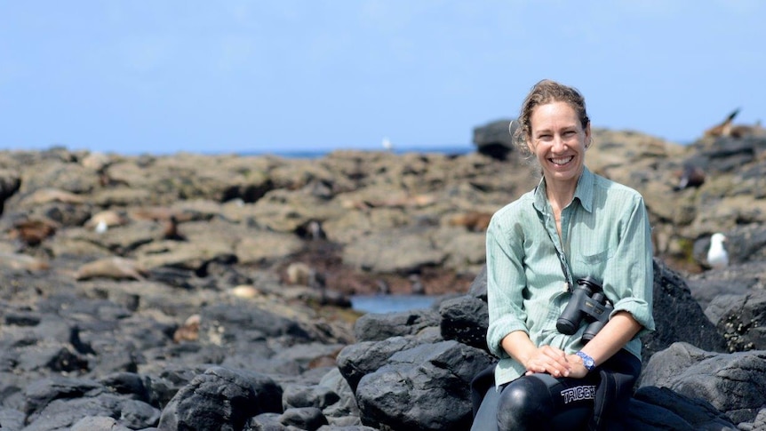 A smiling woman with binoculars around her neck sits on rocks by the ocean.