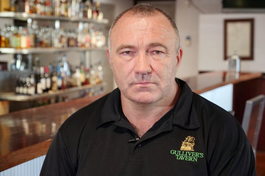 A man wearing a black t-shirt sits in a bar and looks towards the camera.