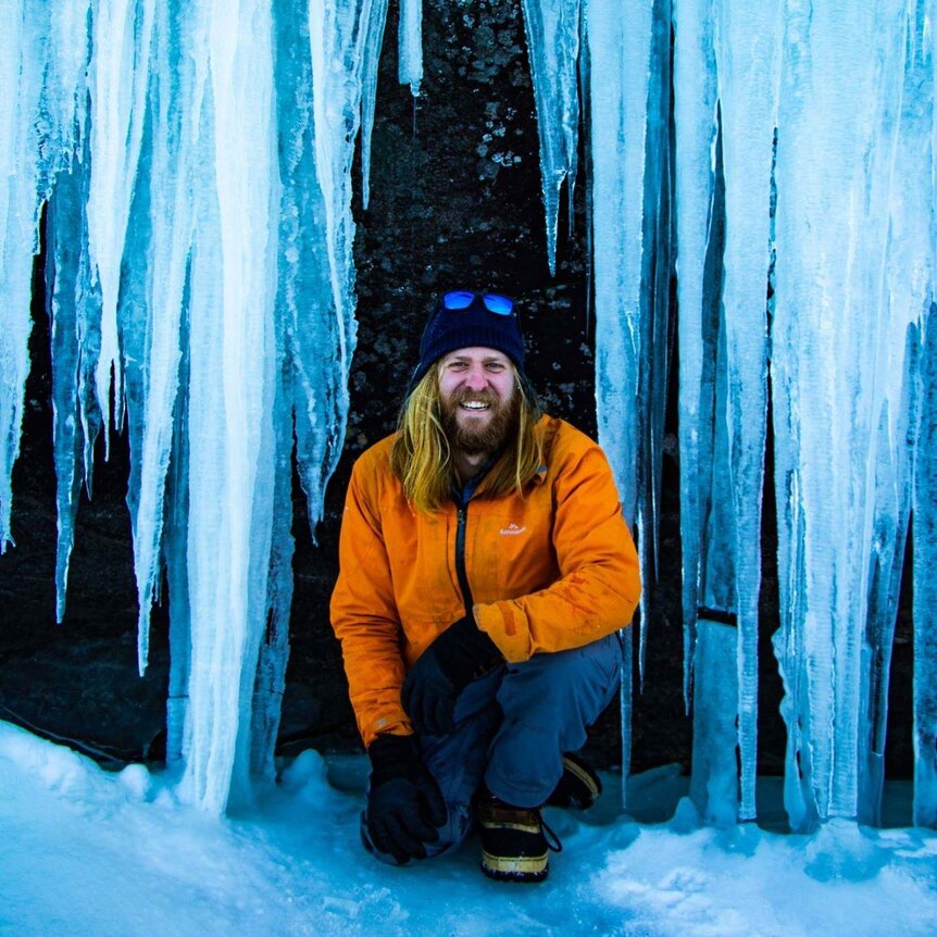 Wilderness guide Luke Brokensha squats in front of huge hanging icicles in the Canadian wilderness