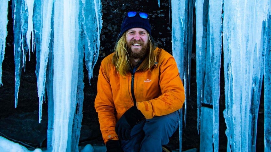 Wilderness guide Luke Brokensha squats in front of huge hanging icicles in the Canadian wilderness