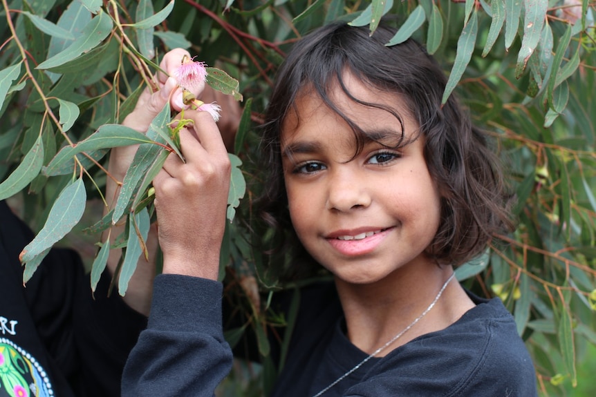 indigenous girl smiling at camera , holding flower of gum leaved tree 