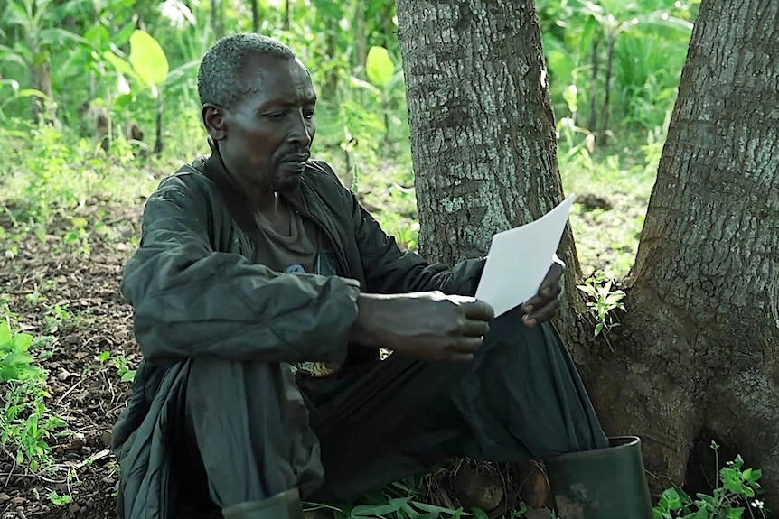 A man sitting, surrounded by trees, looking at a photograph.