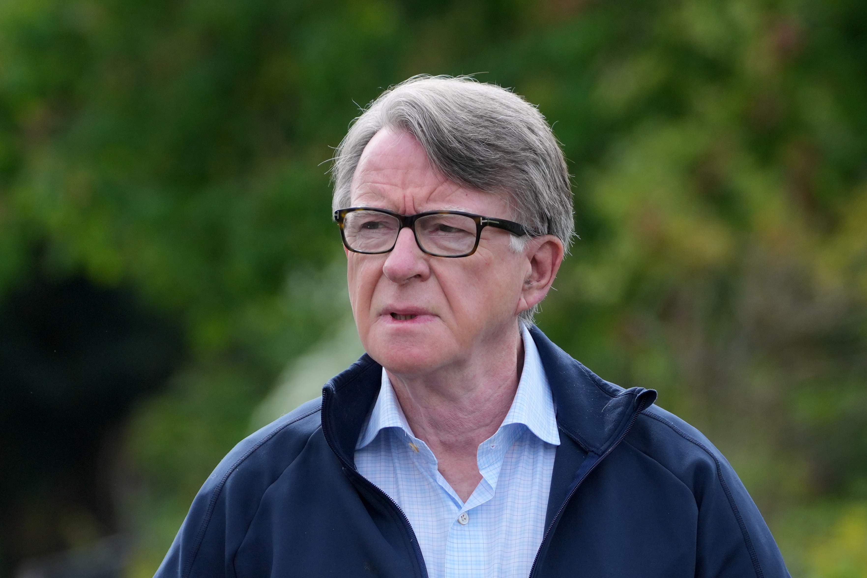 Se ve a un hombre con cabello gris, gafas negras y una camisa abotonada corriendo afuera, con árboles detrás de él.