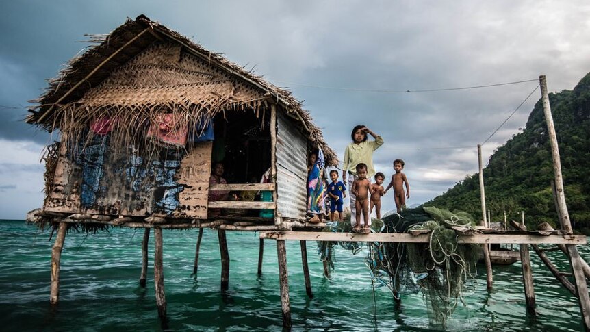 Children stand on the deck of a home on stilts over the water.