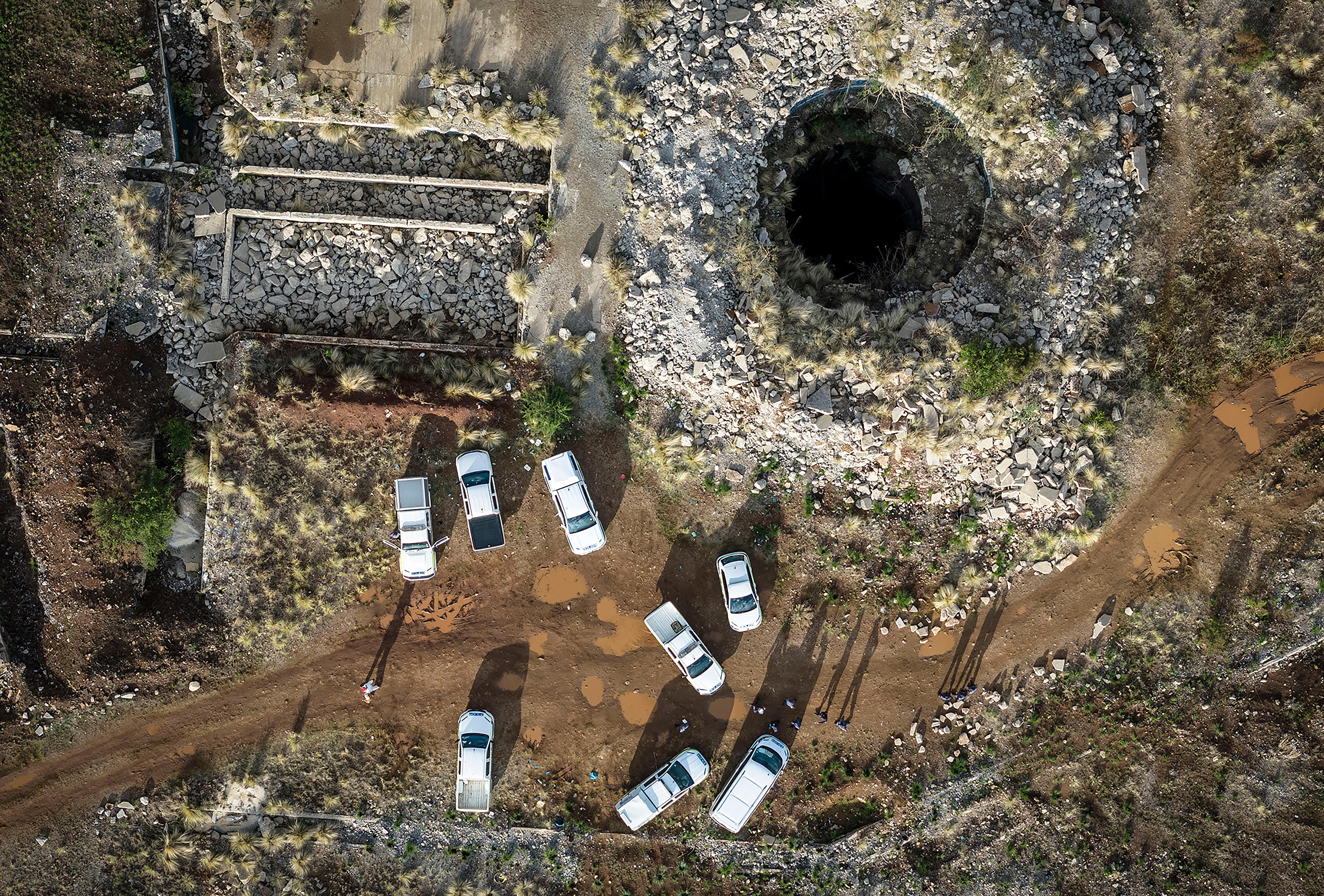 An aerial view of a round mine shaft opening in rocky terrain, with eight white vehivles parked nearby