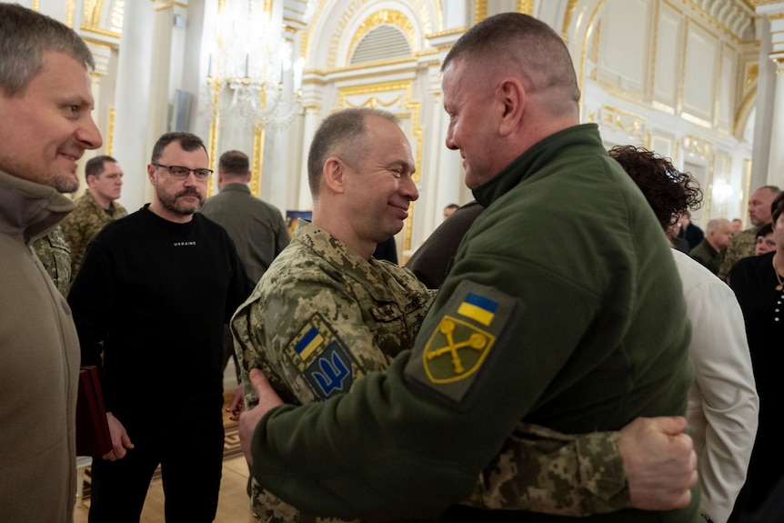 Two men in army uniforms embrace each other among a crowd inside an opulent building.
