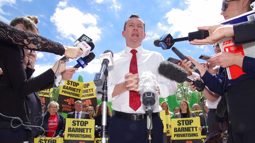 Mark McGowan surrounded by microphones, as anti-privatisation demonstrators hold placards in the background.