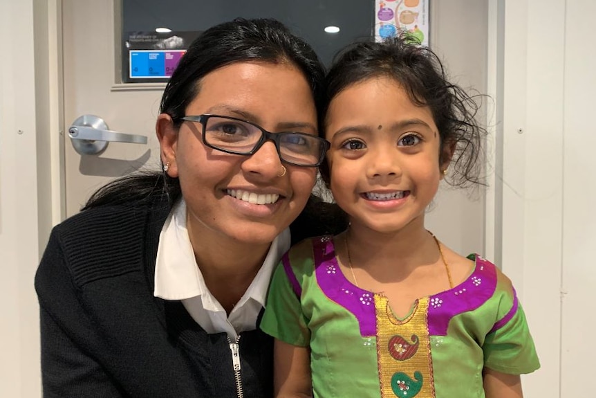 A mum wearing black glasses a white collared business shirt and black jacket crouches beside her smiling daughter.