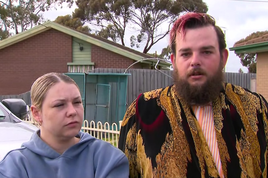 A woman and a man stand outside a suburban home, looking sad.