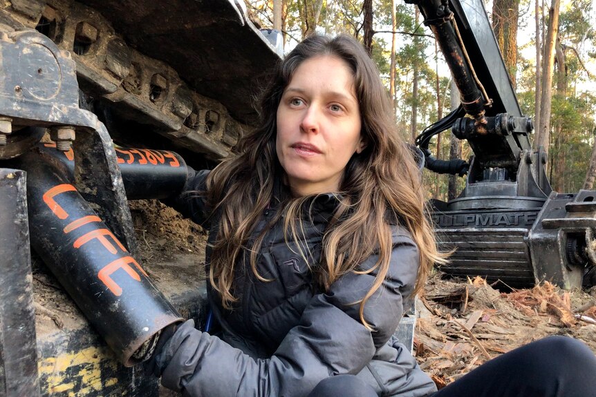 Woman with brown hair locked onto a large logging machine with 'forests' and 'life' written on her arms