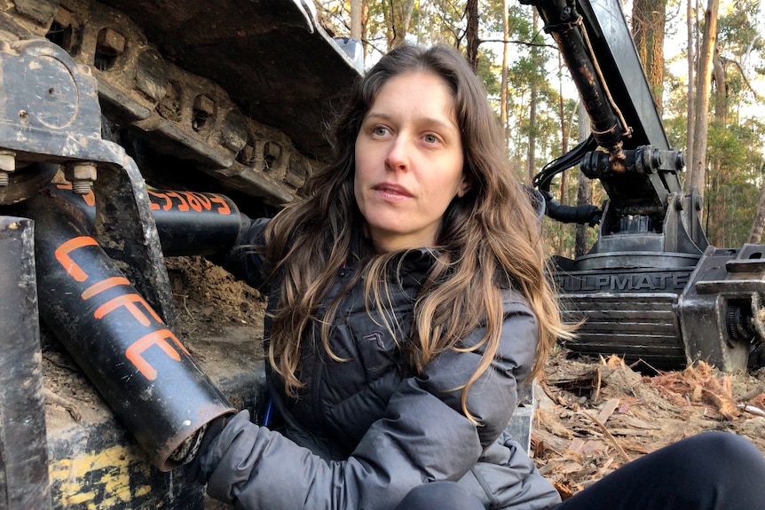 Woman with brown hair locked onto a large logging machine with 'forests' and 'life' written on her arms