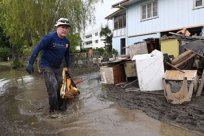 A man walks through knee high muddy water with a house and debris in the background.