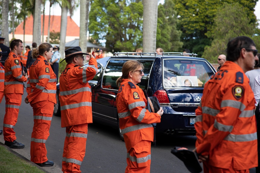 SES volunteers in hi vis orange uniforms line a road where a hearse carrying a coffin travels by, one volunteer salutes