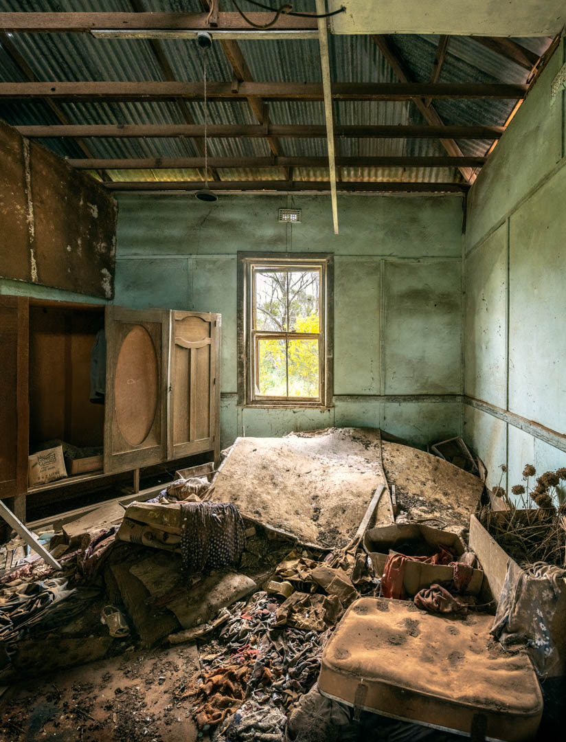 Derelict bedroom with pale blue dirty walls, wooden wardrobes, mattress, blankets on the floor, green outside window.