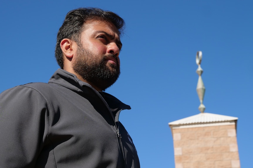 A man with black hair, black moustache and beard in front of a mosque with turret and a iron symbol, blue sky.