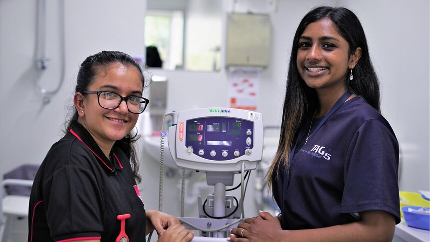 Two teenage female students lean over a piece of medical equipment, both smile broadly to the camera. 