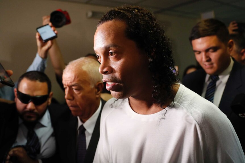 Ronaldinho has his long, curly hair out as he is led through a corridor by a number of people.